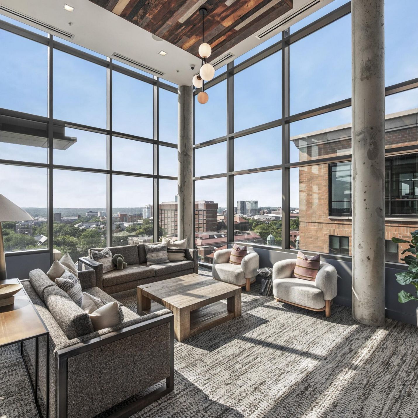 Six11 Student Living Apartments A modern lounge with floor-to-ceiling windows overlooking a city skyline, featuring gray sofas, two armchairs, and a wood coffee table.