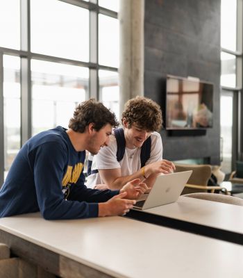 Six11 Student Living Apartments Two young men sit at a table, smiling and looking at a laptop in a bright, modern room with large windows.