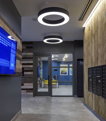 Six11 Student Living Apartments Modern hallway with circular ceiling lights, a wall TV screen, and rows of black mailboxes on the right.