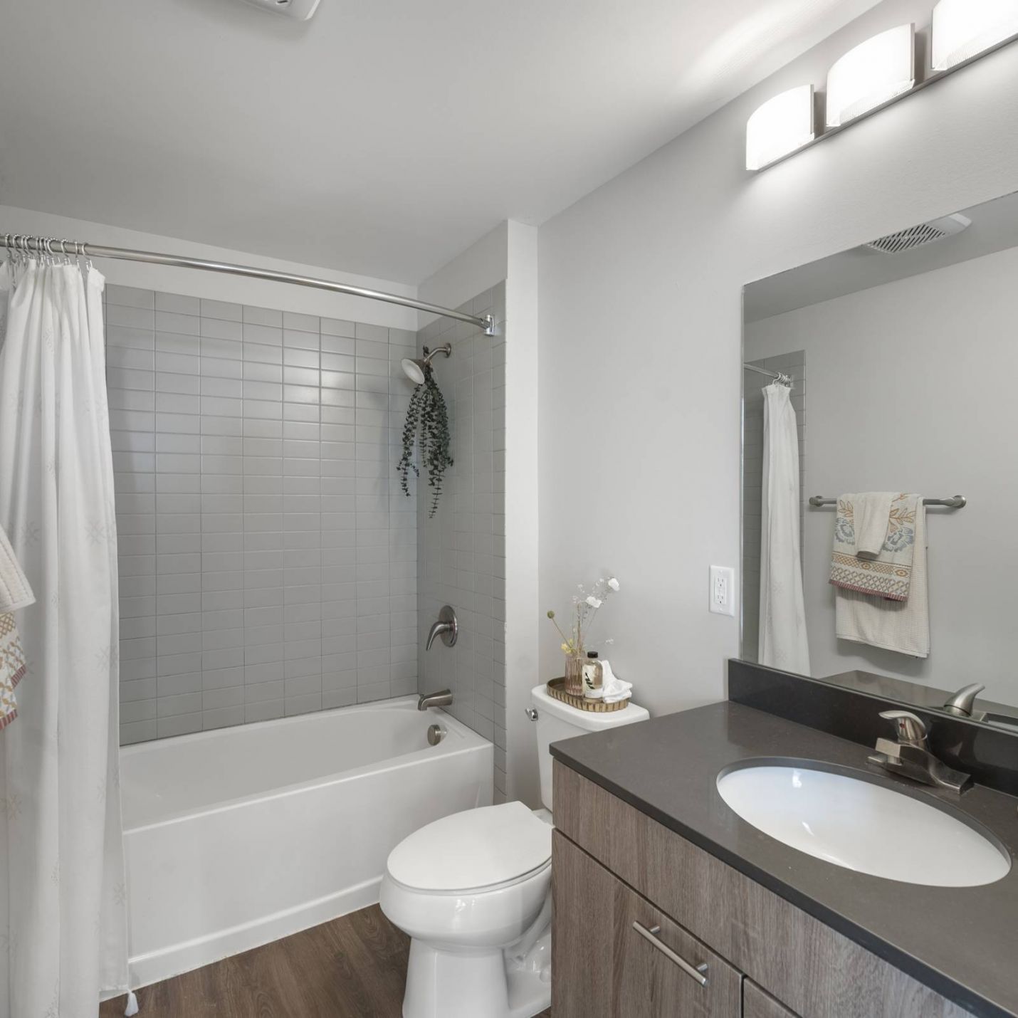Six11 Student Living Apartments Modern bathroom with gray tiled tub, white shower curtain, dark vanity, and lighted mirror.