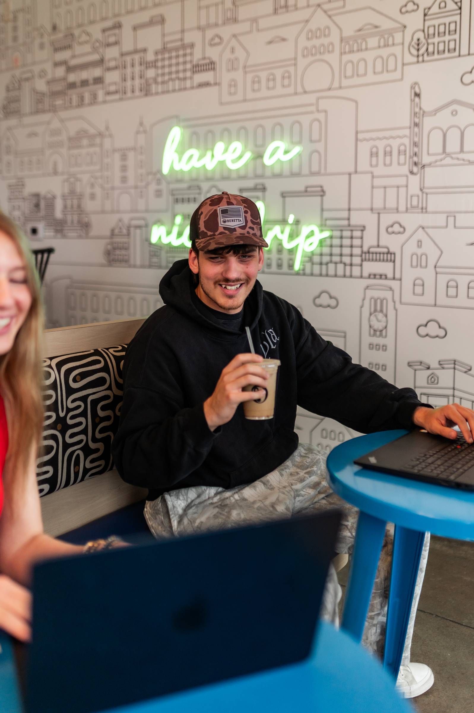 Six11 Student Living Apartments Man in a hoodie and cap smiles while holding a drink and using a laptop in a café with neon wall art.