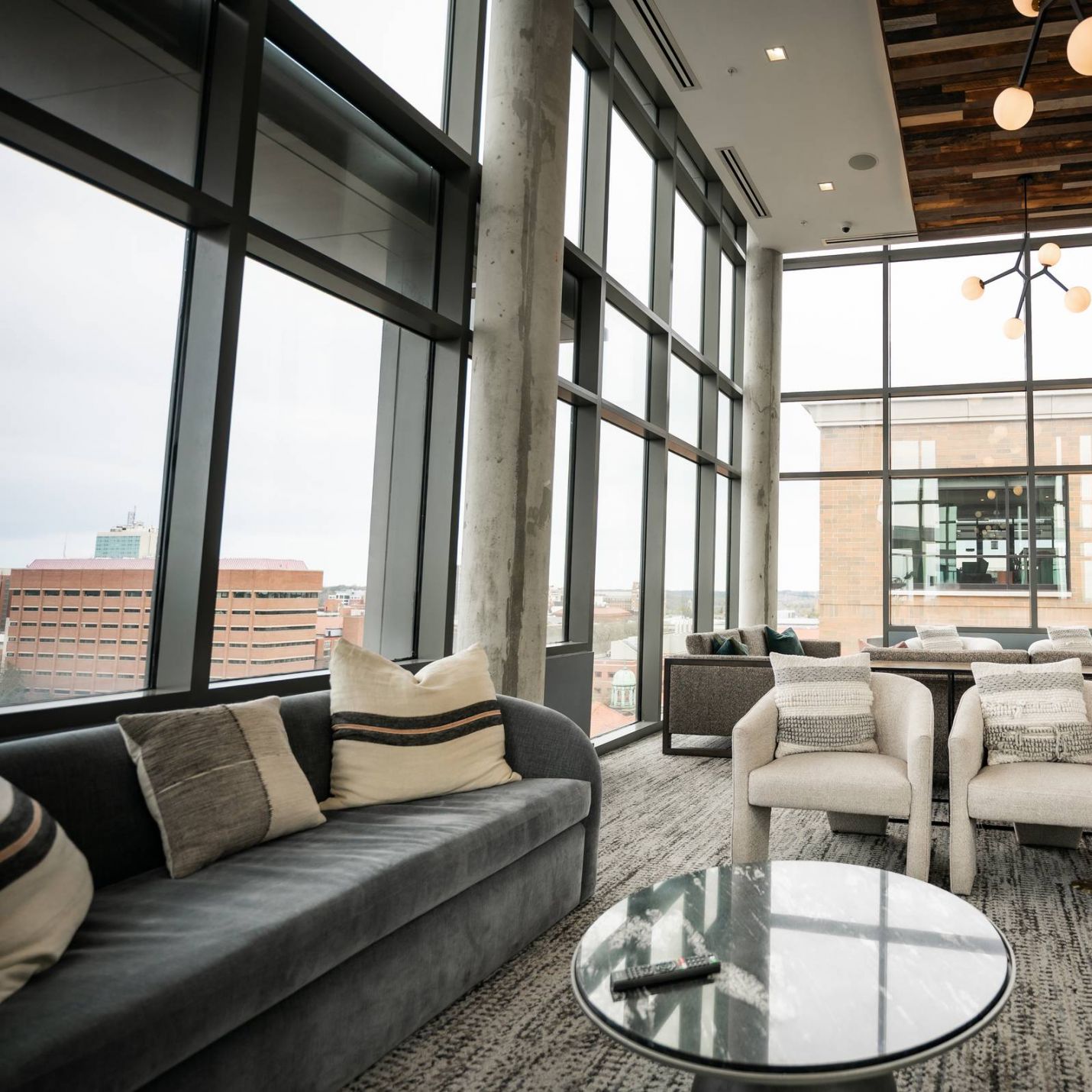 Six11 Student Living Apartments Modern lounge with large windows, grey sofas, armchairs, a round glass table, and city view outside.