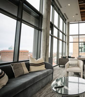 Six11 Student Living Apartments Modern lounge with large windows, grey sofas, armchairs, a round glass table, and city view outside.