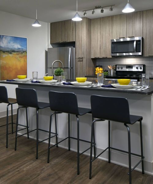 Six11 Student Living Apartments Modern kitchen with wood cabinets, stainless steel appliances, island seating, and colorful table settings.