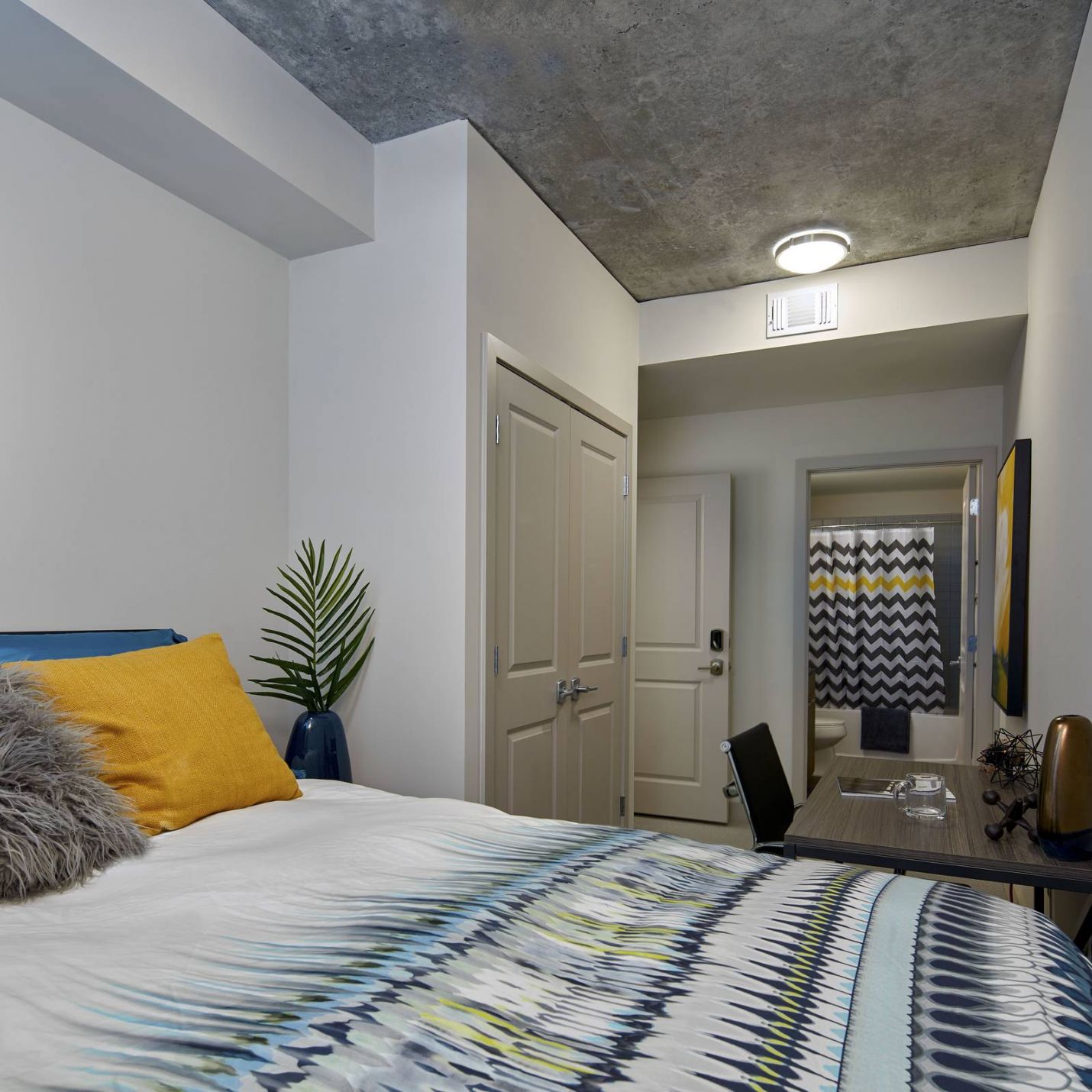 Six11 Student Living Apartments Modern bedroom with abstract art, patterned bedding, desk, and a plant, leading to a bathroom with zigzag shower curtain.