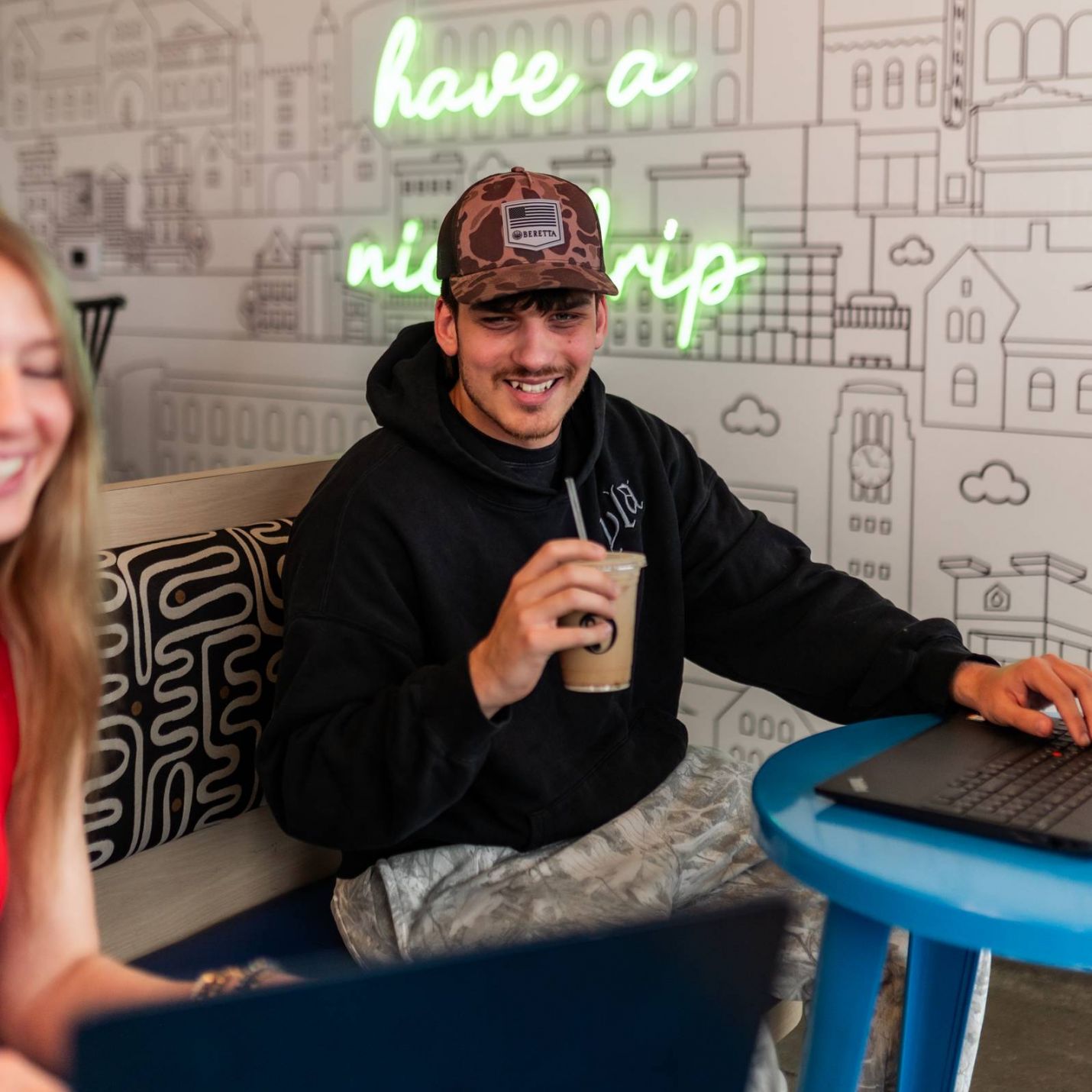 Six11 Student Living Apartments Man in a hoodie and cap smiles while holding a drink and using a laptop in a café with neon wall art.